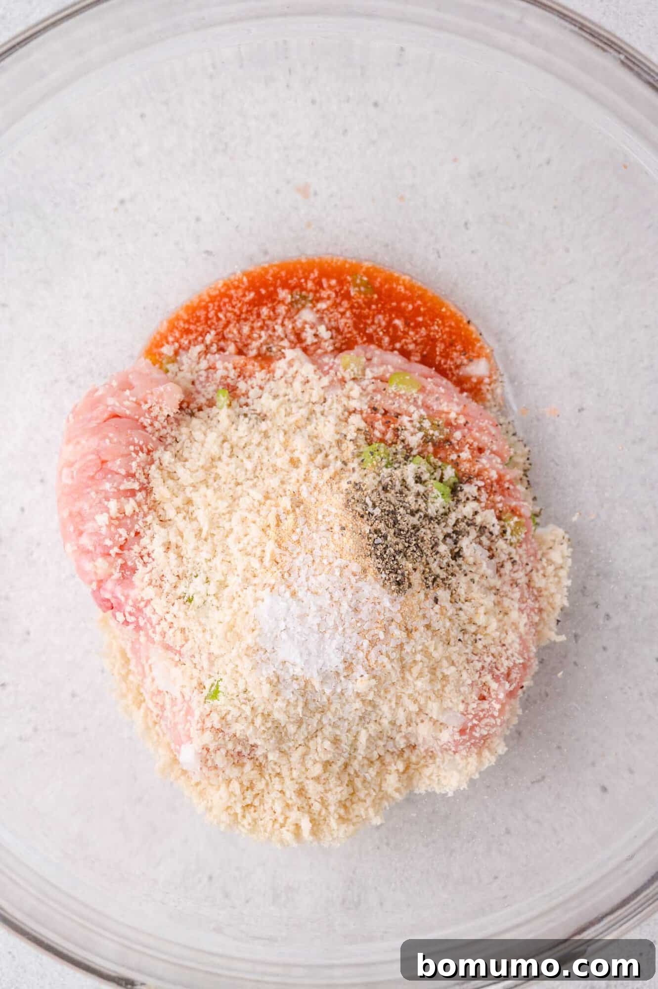 Ground meat, breadcrumbs, seasoning, and sauce in a glass mixing bowl, ready to be combined for a recipe.