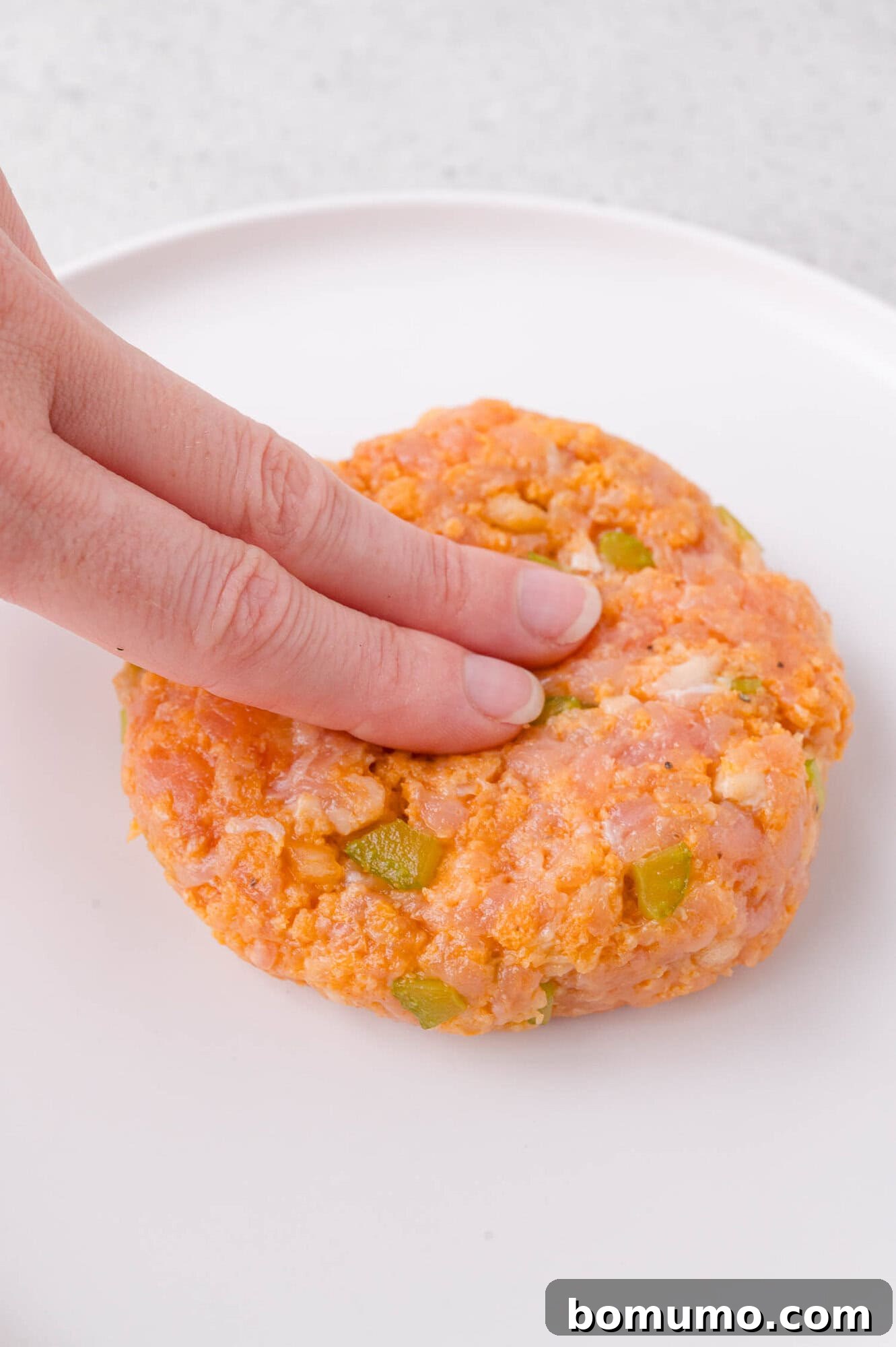 A hand presses down on a raw chicken burger mixed with chopped vegetables on a white plate.
