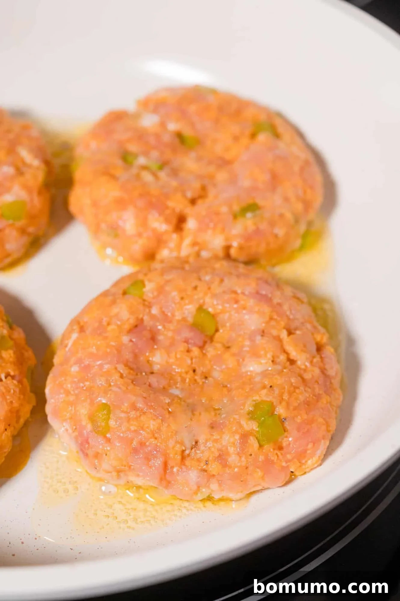 Raw seasoned ground chicken patties with visible celery pieces are arranged in a white skillet with some oil.