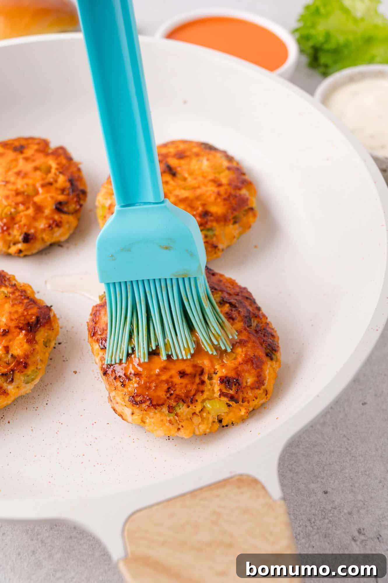 A blue silicone brush applies sauce to a cooked patty in a white skillet, with three other patties and bowls of sauces in the background.