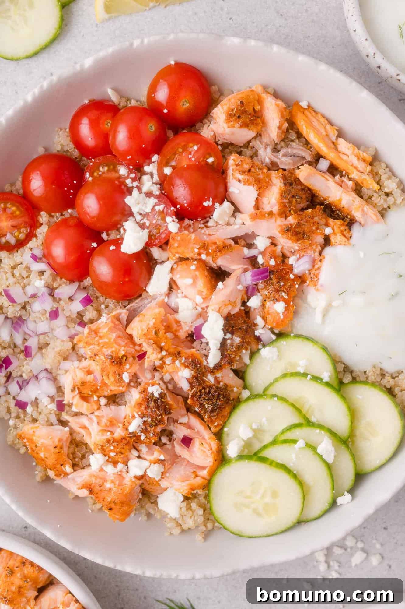 A bowl with quinoa, flaked salmon, cherry tomatoes, cucumber slices, red onion, crumbled feta, and a dollop of white sauce.