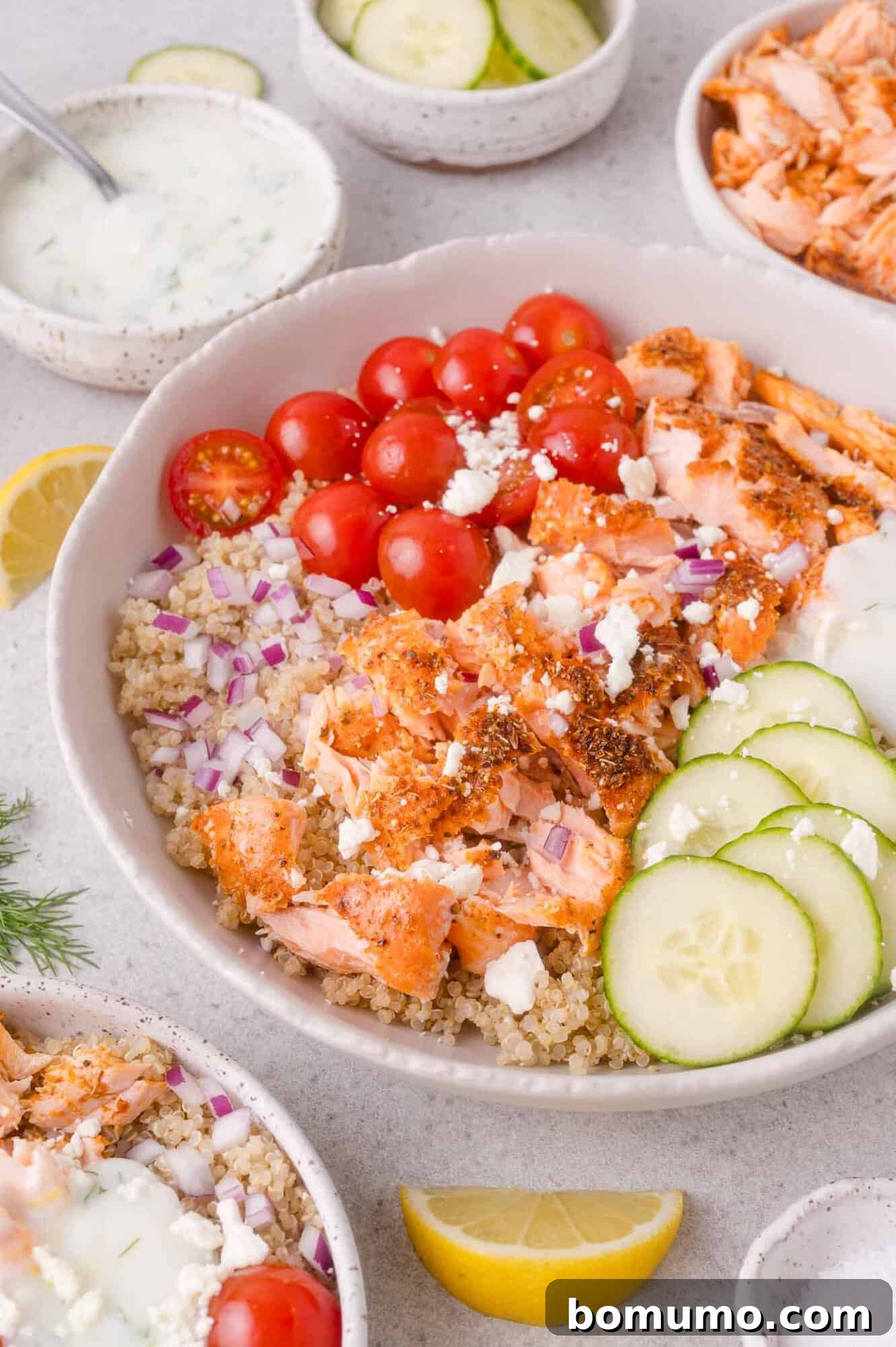 Bowl with quinoa, sliced cucumbers, cherry tomatoes, red onions, crumbled feta, and seasoned salmon, served with a side of creamy dressing and lemon wedges.