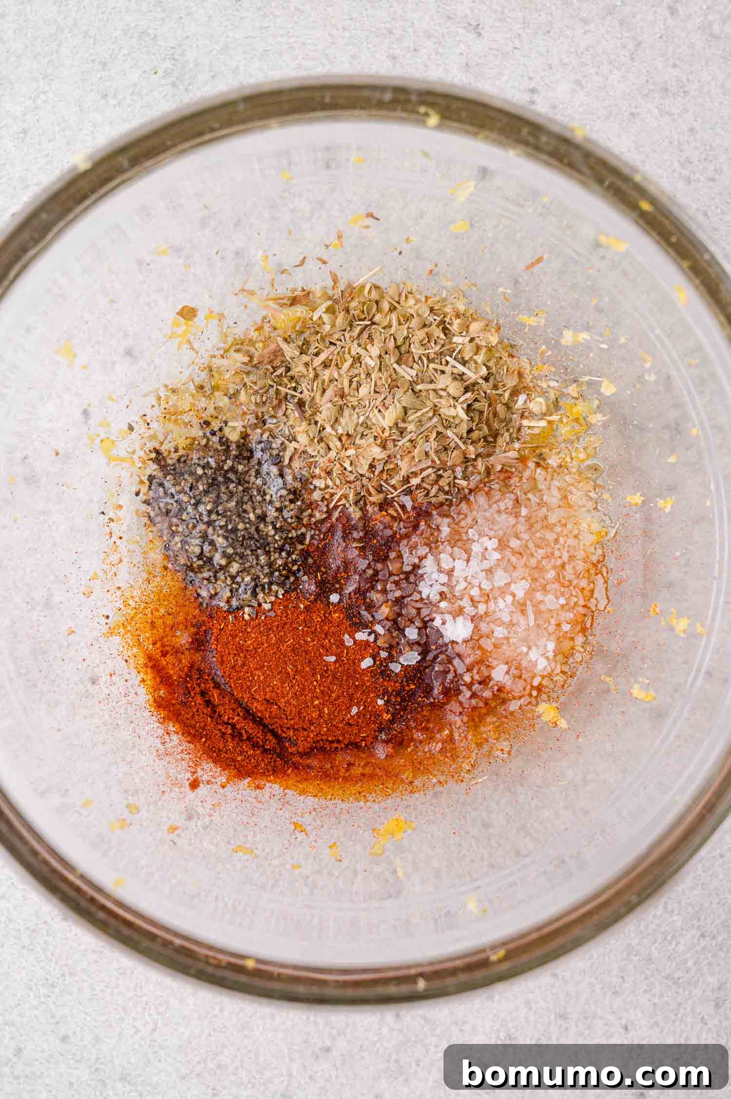 A glass bowl containing piles of dried oregano, ground black pepper, salt, paprika, and lemon zest on a light surface.