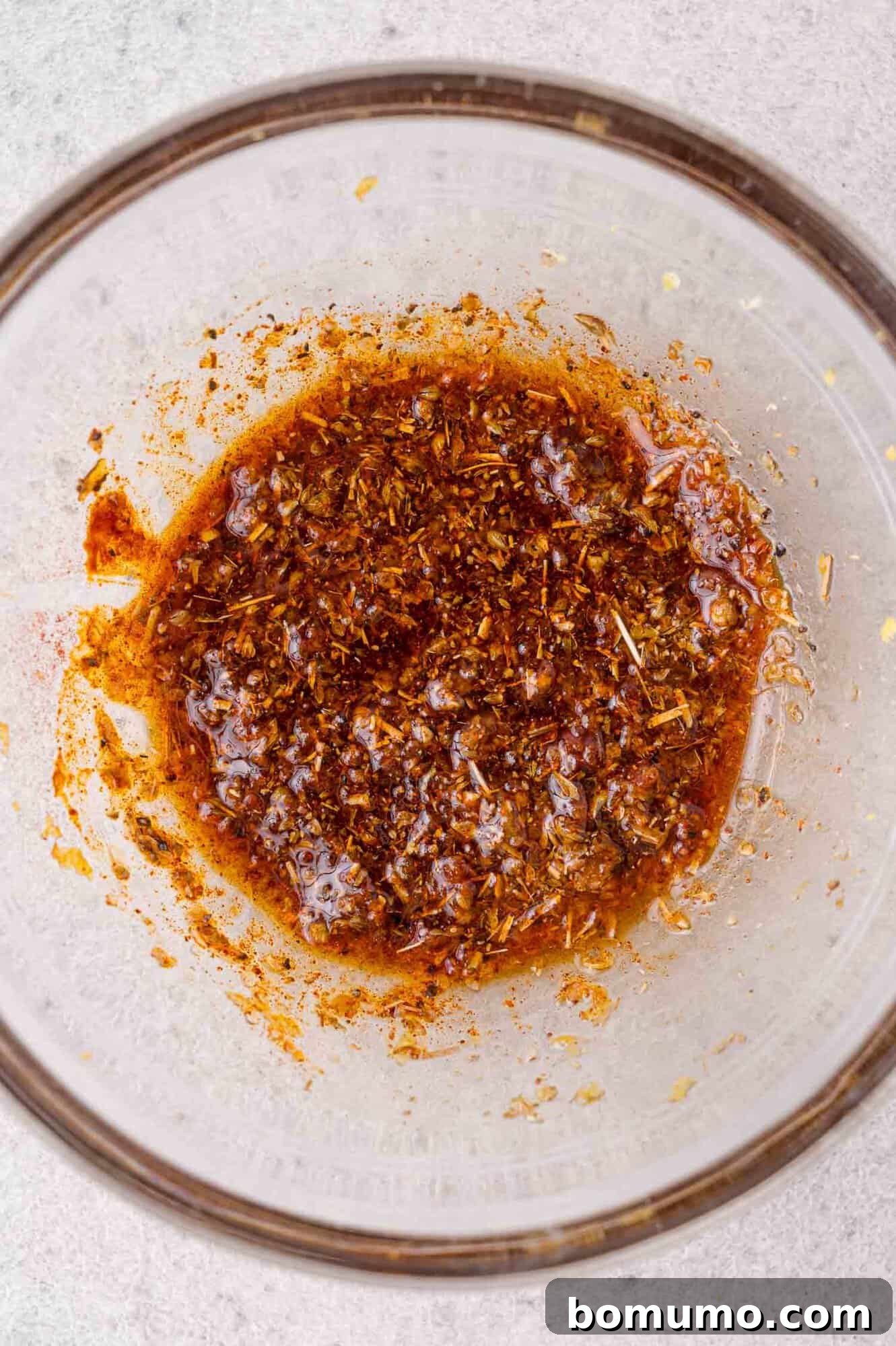 A glass bowl containing a reddish-brown spice marinade with visible herbs and oil, placed on a light surface.