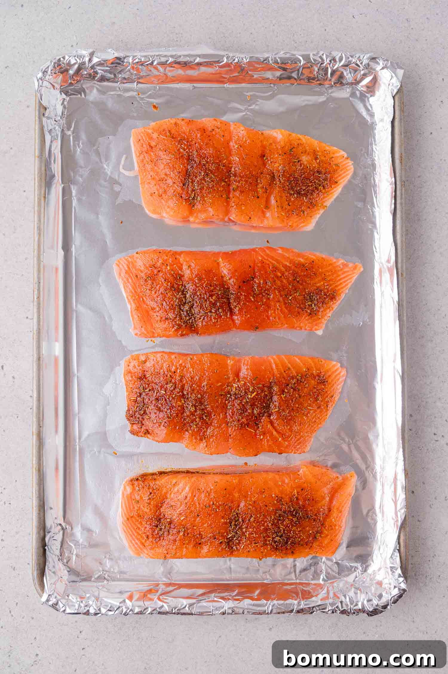 Four raw salmon fillets with seasoning are arranged in a row on a foil-lined baking sheet.