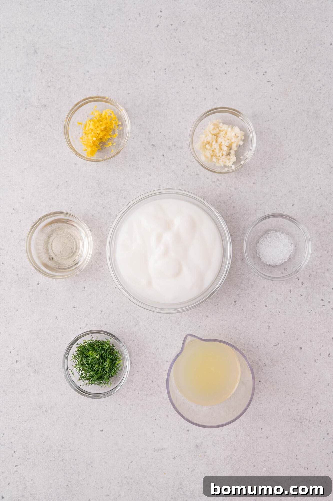 Six small bowls containing yogurt, chopped dill, minced garlic, lemon zest, salt, lemon juice, and vinegar arranged on a light-colored surface.