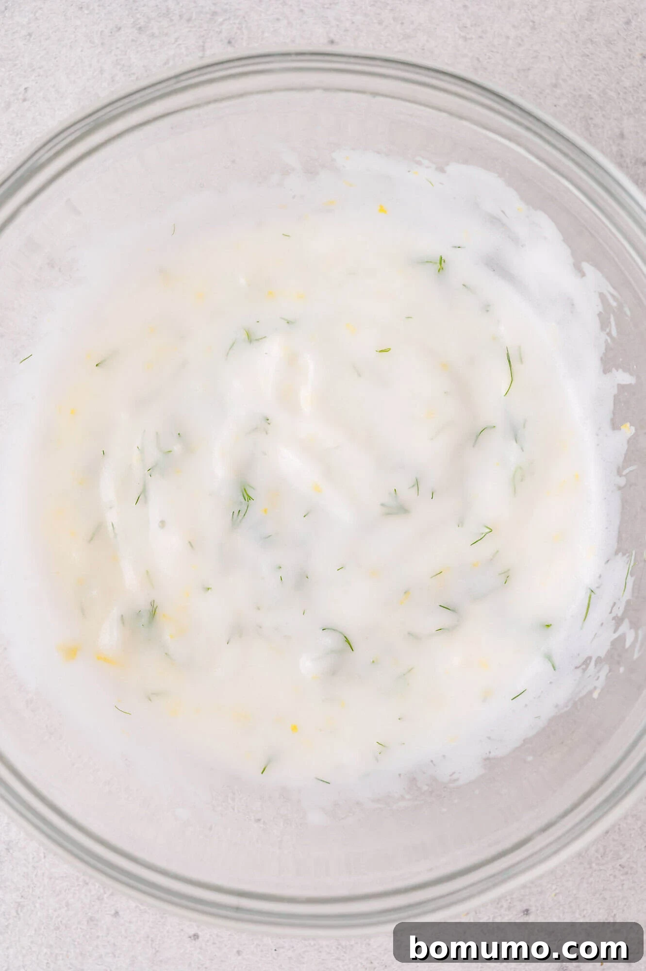 Clear glass bowl containing a white, creamy sauce with dill mixed in, set on a light-colored surface.