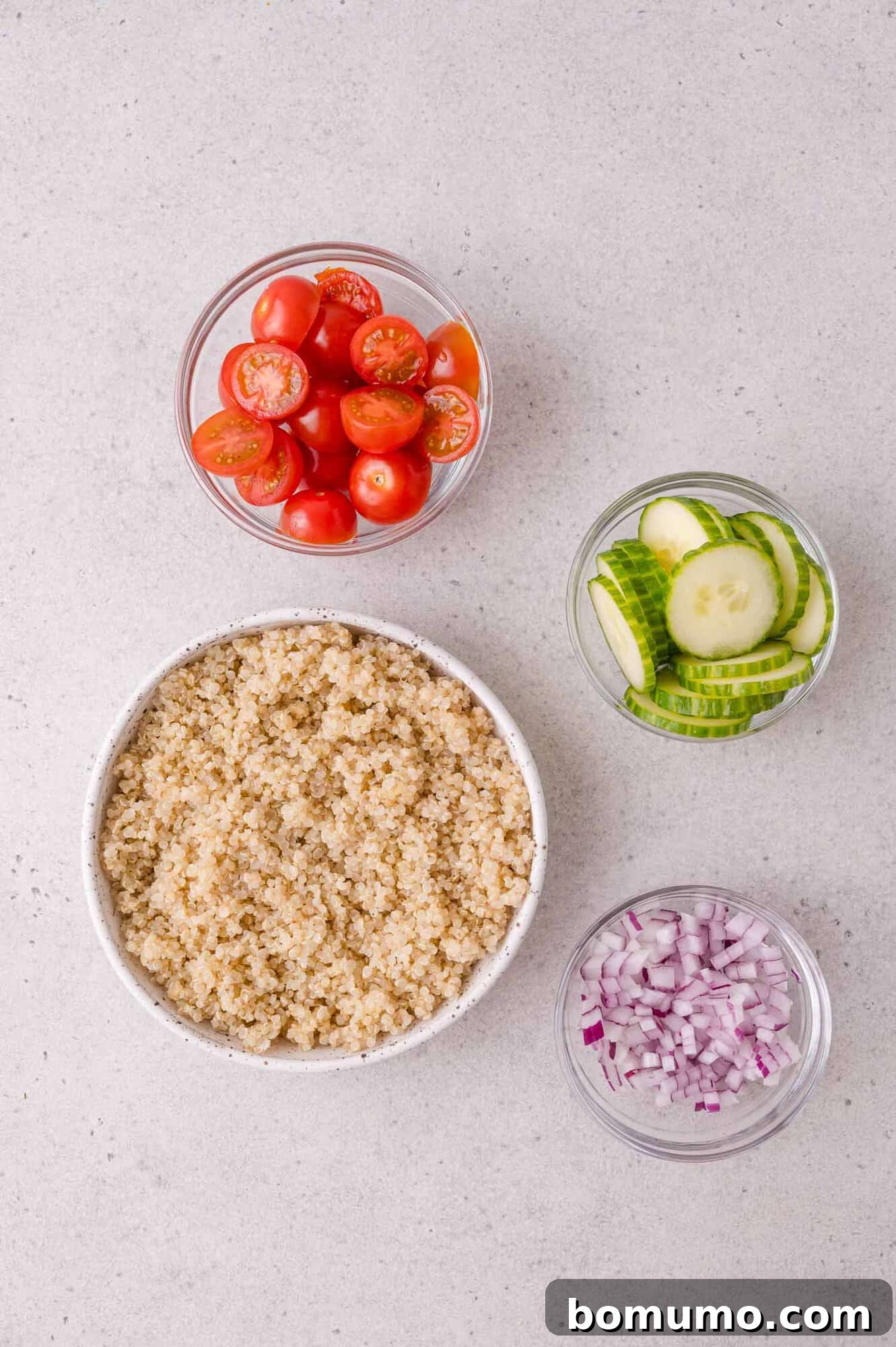 A bowl of cooked quinoa, and three smaller bowls containing halved cherry tomatoes, sliced cucumbers, and diced red onions, all on a light countertop.