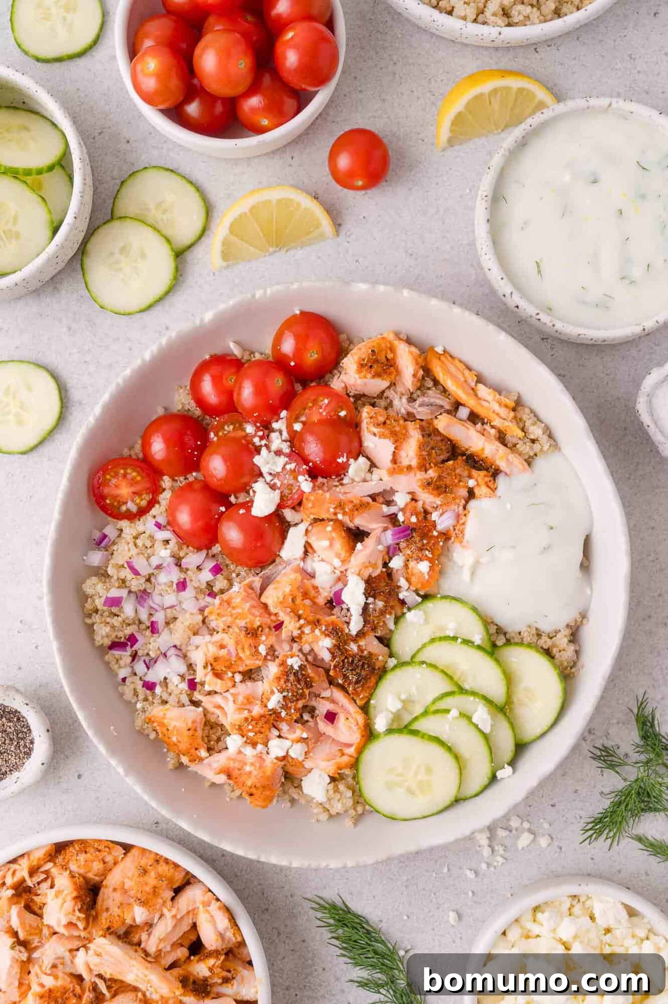 Bowl with quinoa, cooked salmon, cherry tomatoes, cucumber slices, red onion, feta, and tzatziki sauce. Surrounded by small bowls of ingredients and lemon wedges on a white background.