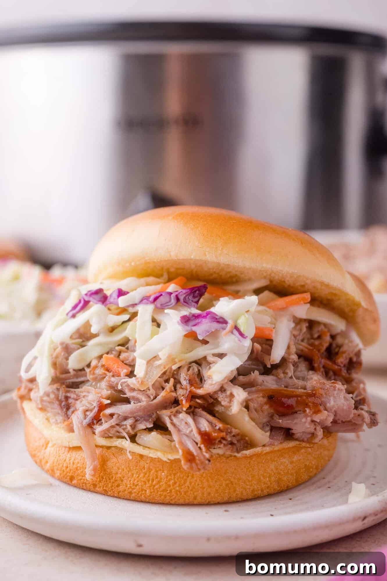 Slow Cooker Apple Cider Pulled Pork Sandwiches 2 Pulled pork sandwich topped with coleslaw on a white bun, served on a plate with a slow cooker in the background.