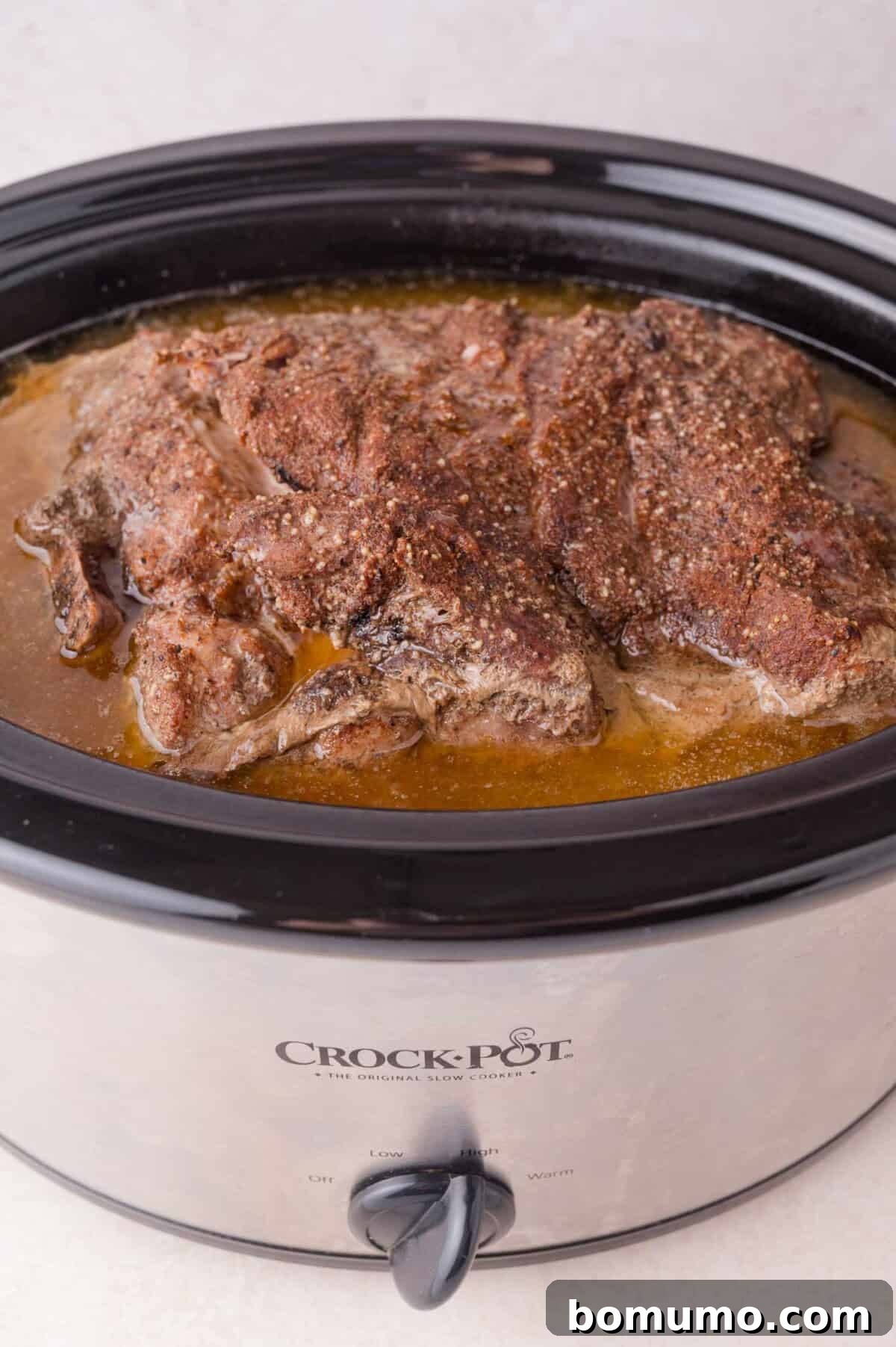 Slow Cooker Apple Cider Pulled Pork Sandwiches 8 Cooked seasoned pork sits in broth inside a black Crock-Pot slow cooker on a light countertop.