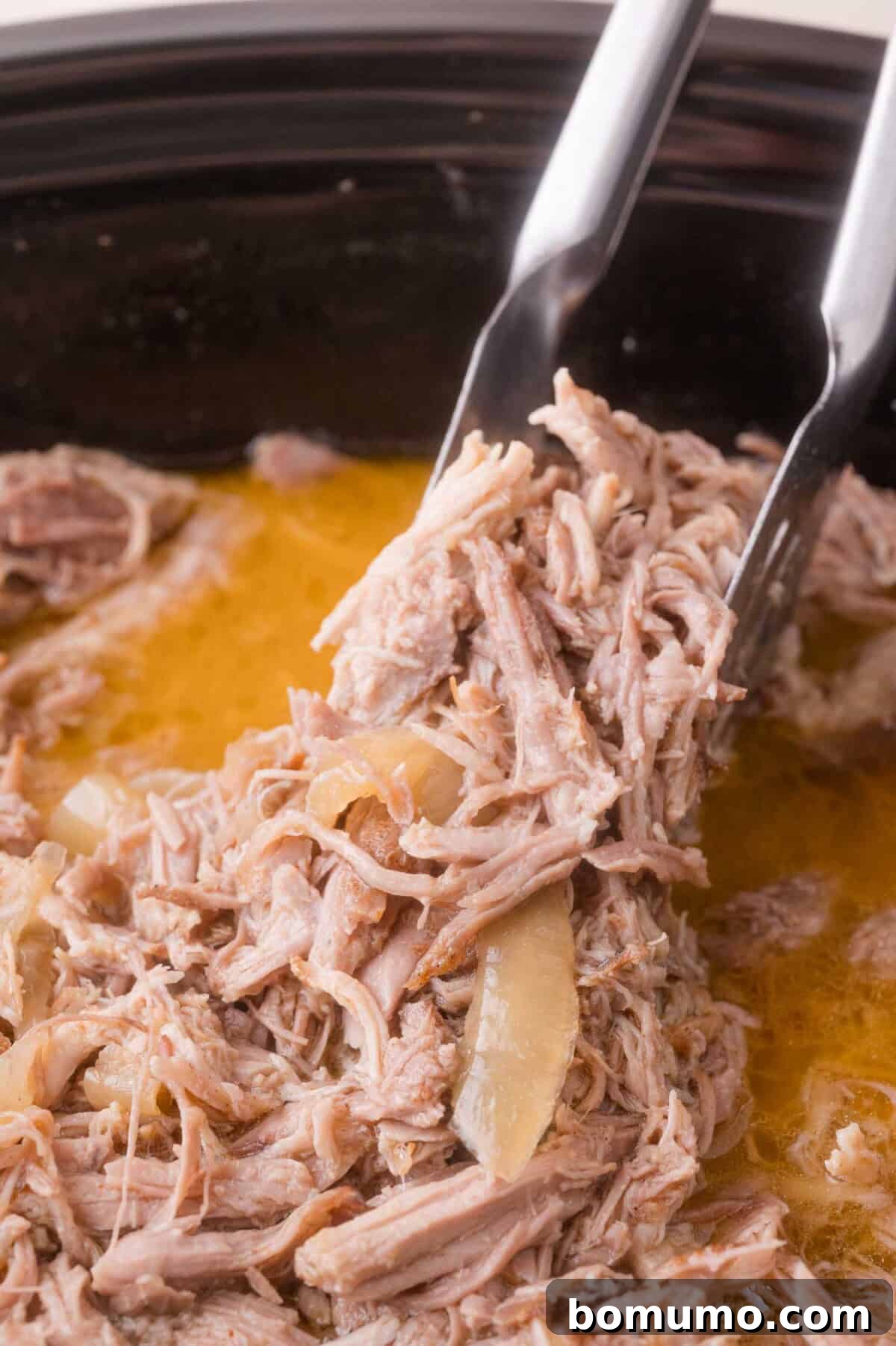 Slow Cooker Apple Cider Pulled Pork Sandwiches 10 Cooked shredded pork being lifted from a slow cooker with metal tongs, surrounded by broth and pieces of onion.