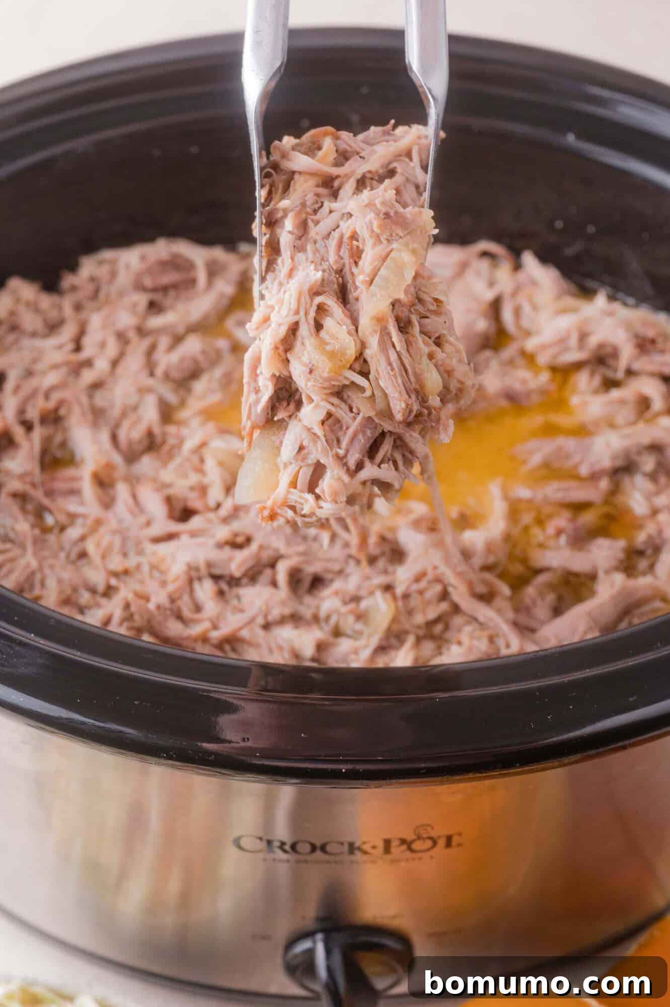 Slow Cooker Apple Cider Pulled Pork Sandwiches 3 Shredded pork being lifted with tongs from a slow cooker filled with cooked meat and juices.