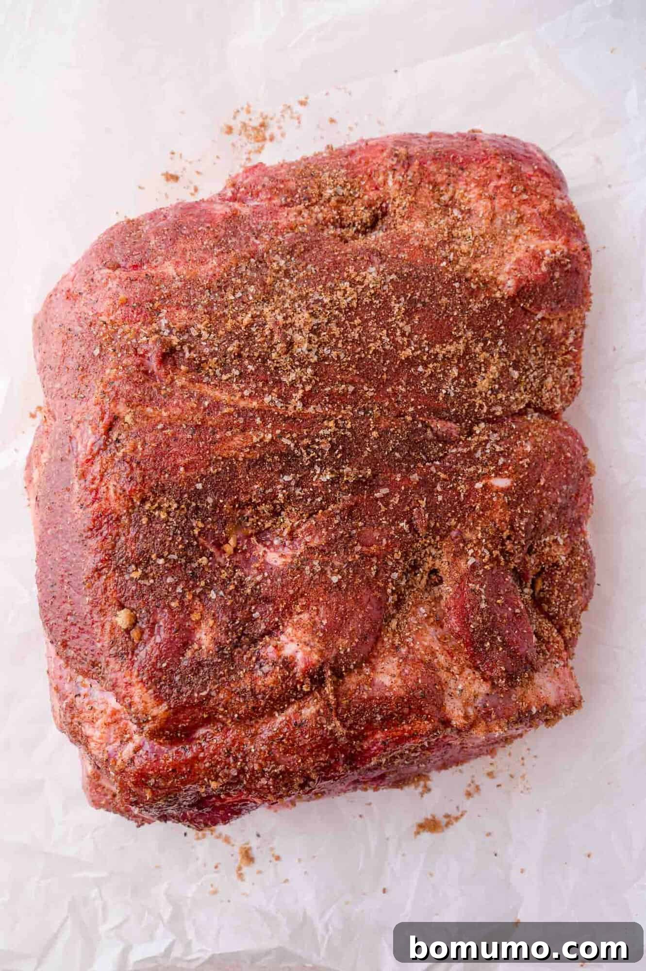 Slow Cooker Apple Cider Pulled Pork Sandwiches 7 A raw cut of pork covered with a dry spice rub sits on a sheet of white parchment paper.
