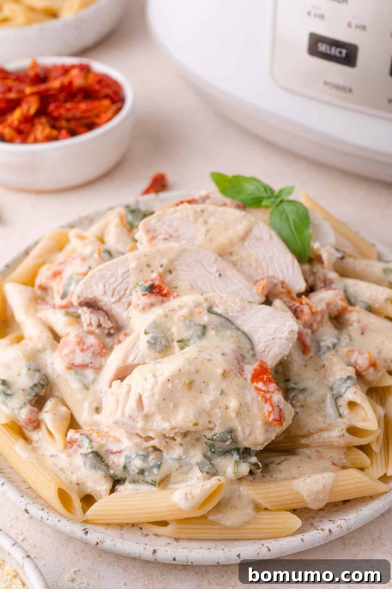 Creamy Tuscan Chicken in the Slow Cooker 2 A plate of penne pasta topped with creamy sauce, sliced cooked chicken breast, sun-dried tomatoes, and fresh basil.