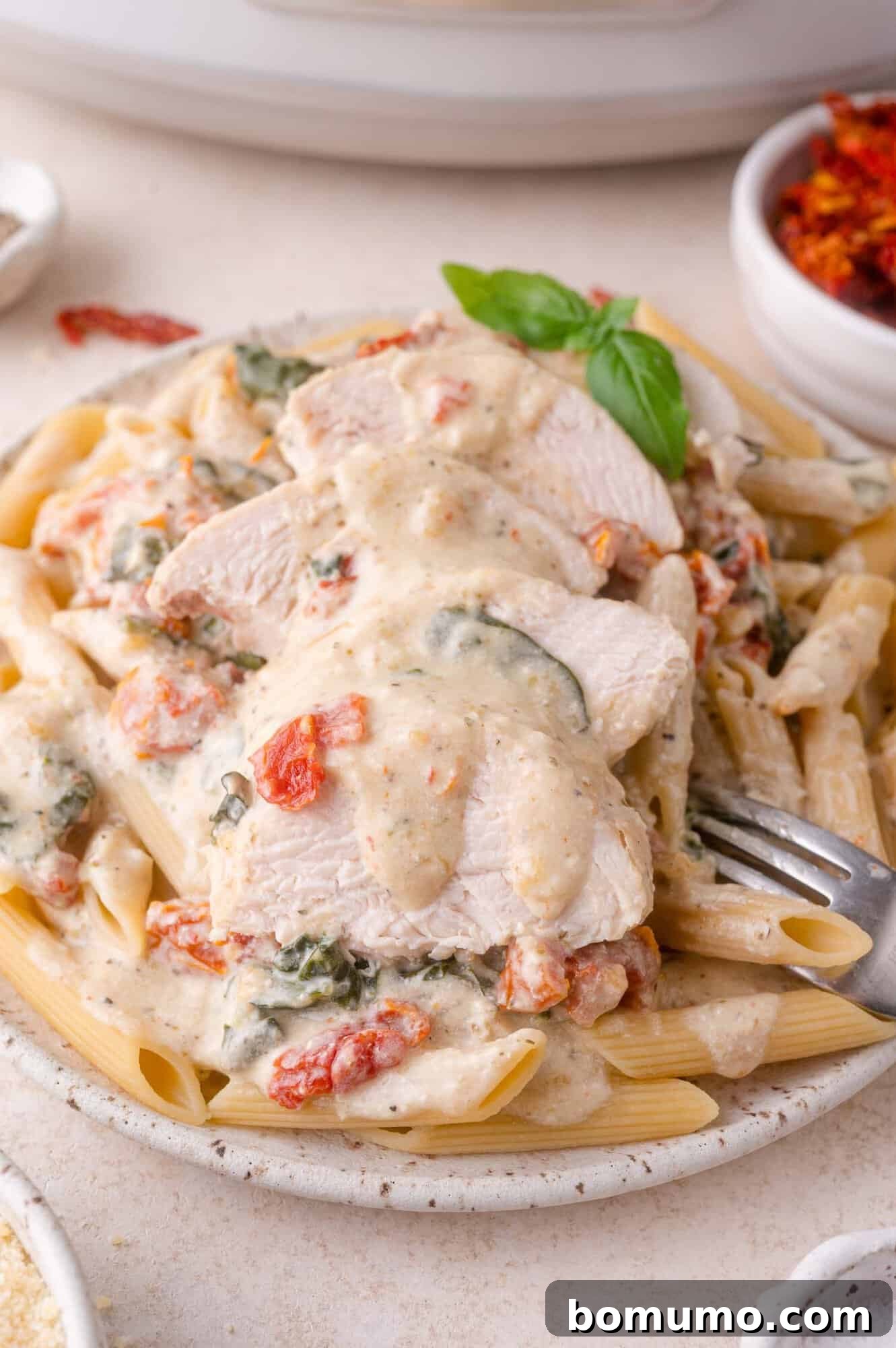 Creamy Tuscan Chicken in the Slow Cooker 8 A plate of penne pasta topped with creamy sauce, sliced chicken breast, sun-dried tomatoes, and spinach, garnished with a basil leaf.