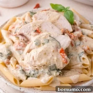 Creamy Tuscan Chicken in the Slow Cooker 9 Plate of penne pasta topped with sliced chicken breast, creamy white sauce, spinach, and sun-dried tomatoes, garnished with a basil sprig.