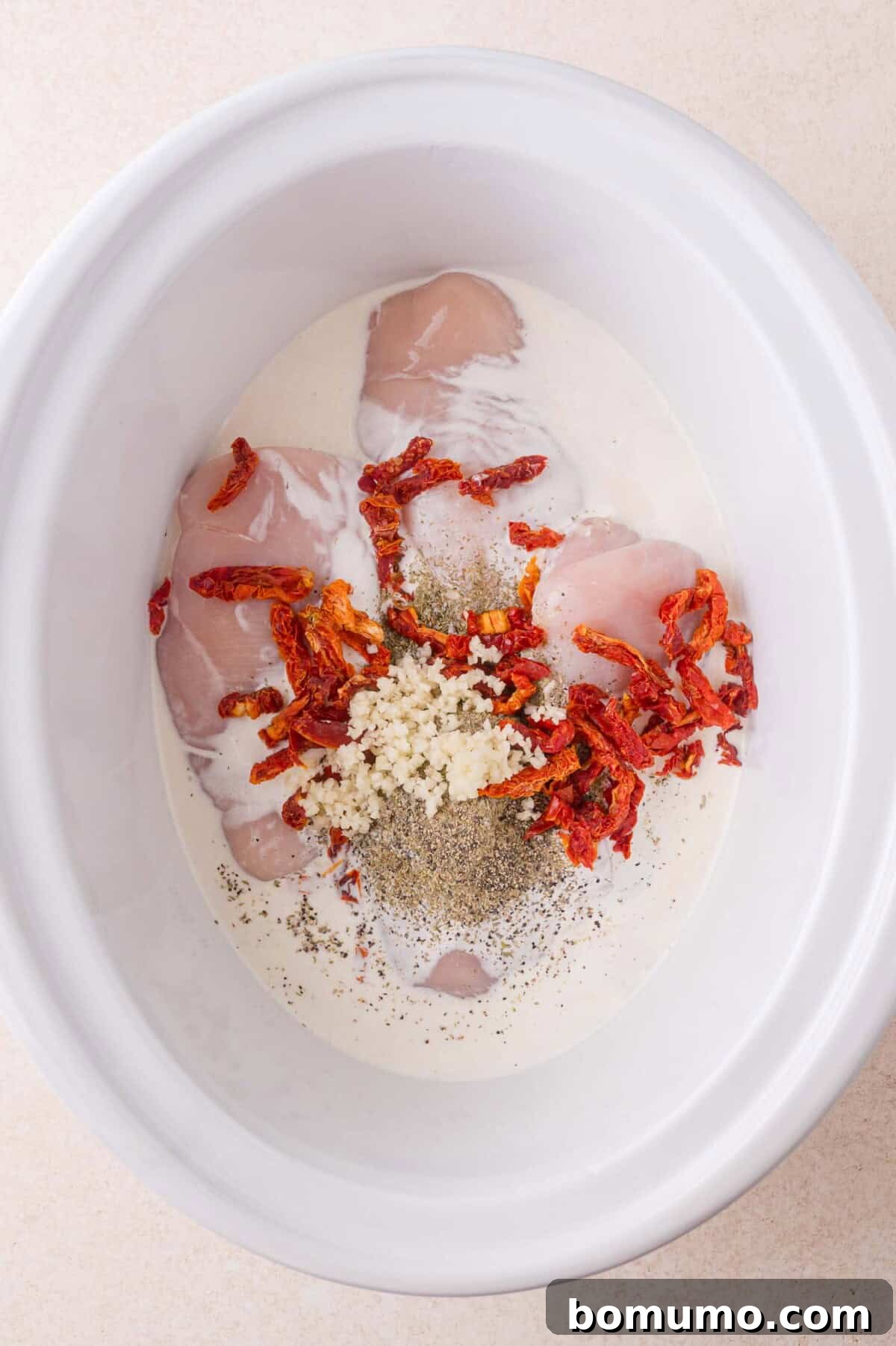 Creamy Tuscan Chicken in the Slow Cooker 5 Raw chicken breasts, cream, sun-dried tomatoes, minced garlic, and black pepper are placed in a white slow cooker before mixing.