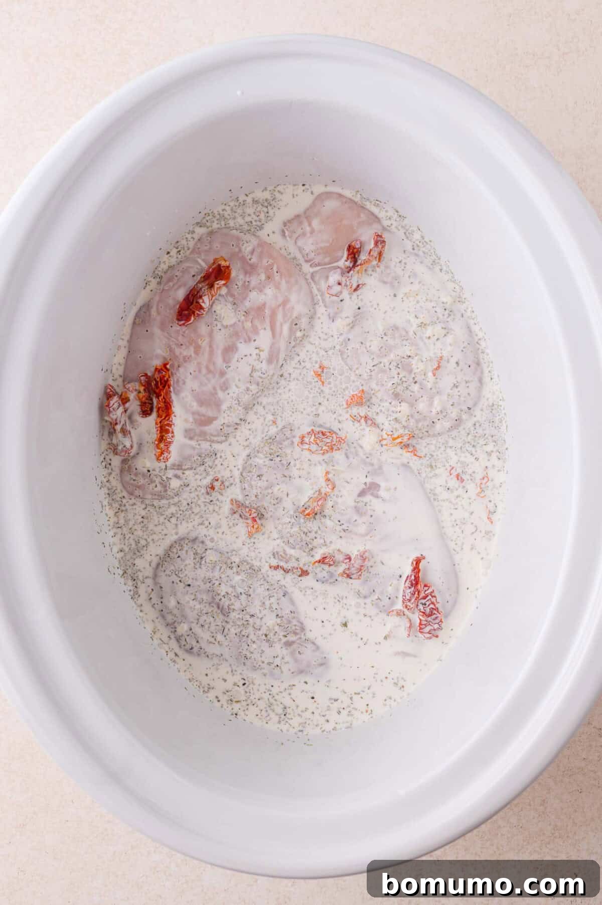 Creamy Tuscan Chicken in the Slow Cooker 6 Raw chicken breasts in a white slow cooker with cream, black pepper, and sun-dried tomato pieces.
