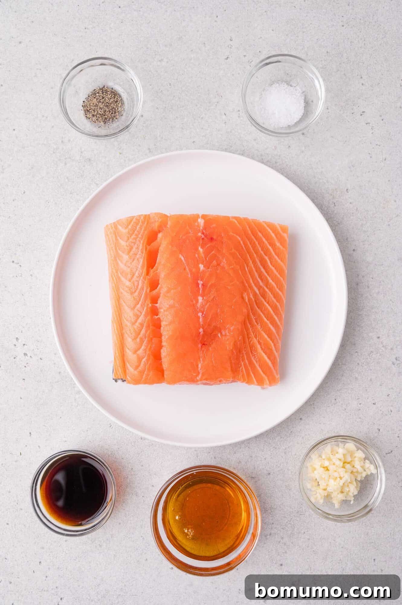 Crispy Honey Garlic Salmon Bites in the Air Fryer 3 Raw salmon fillets on a white plate, surrounded by bowls of soy sauce, honey, minced garlic, black pepper, and salt on a light gray surface.