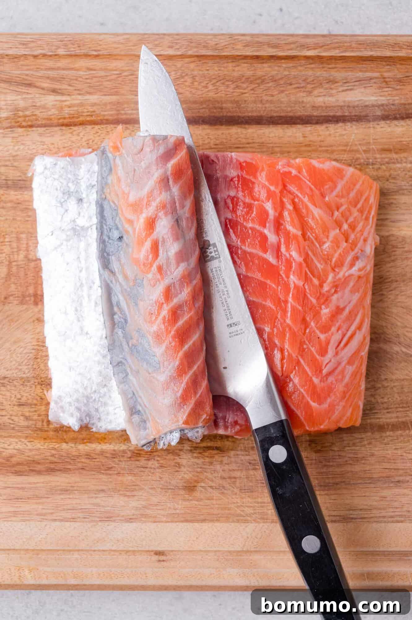 Crispy Honey Garlic Salmon Bites in the Air Fryer 4 A knife is being used to separate the skin from a raw salmon fillet on a wooden cutting board.
