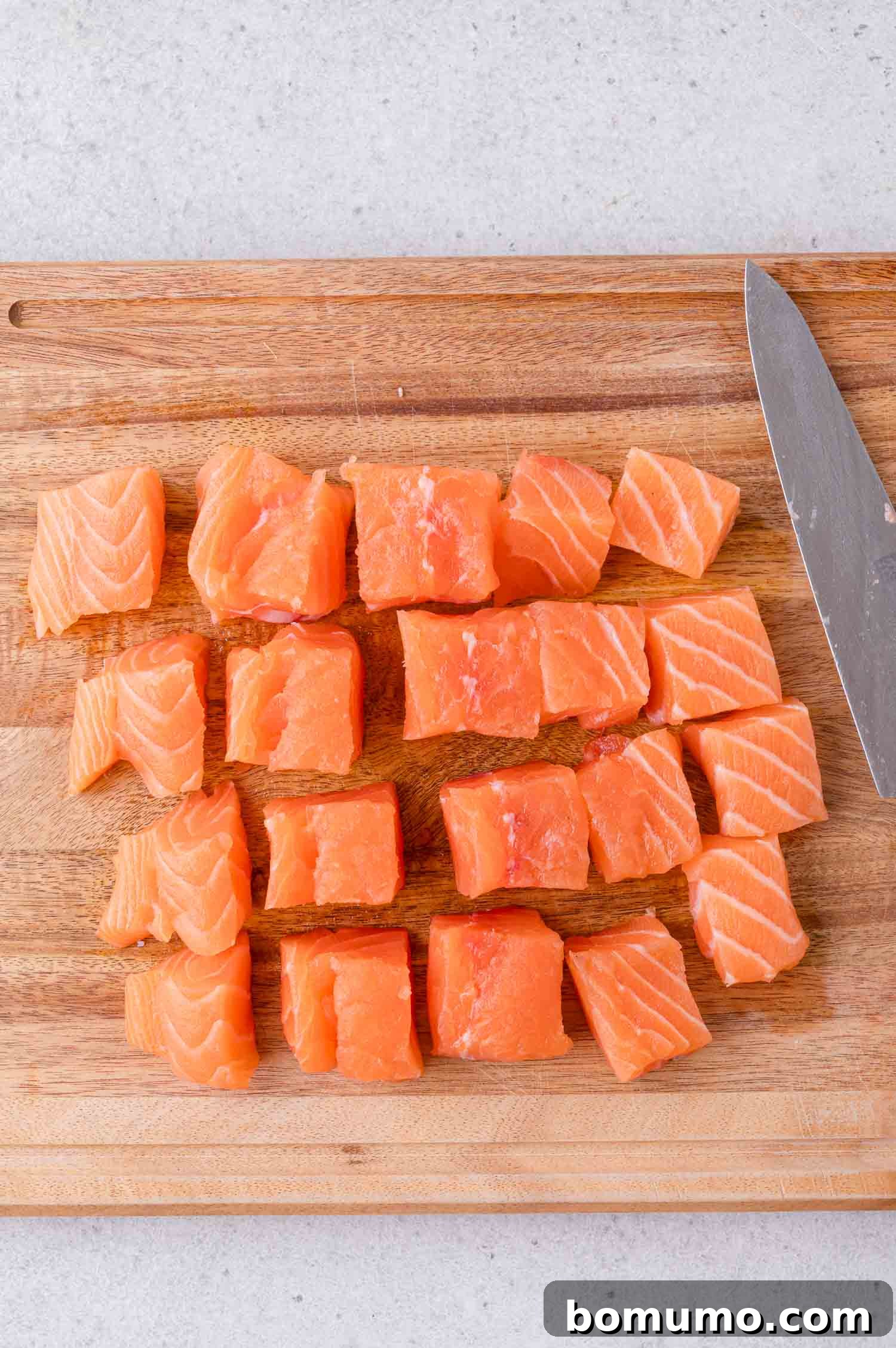 Crispy Honey Garlic Salmon Bites in the Air Fryer 5 Cubed pieces of raw salmon are arranged in rows on a wooden cutting board next to a knife.