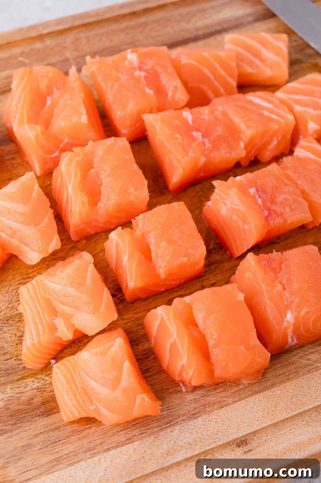 Crispy Honey Garlic Salmon Bites in the Air Fryer 6 Cubes of raw salmon arranged on a wooden cutting board with a knife partially visible in the corner.