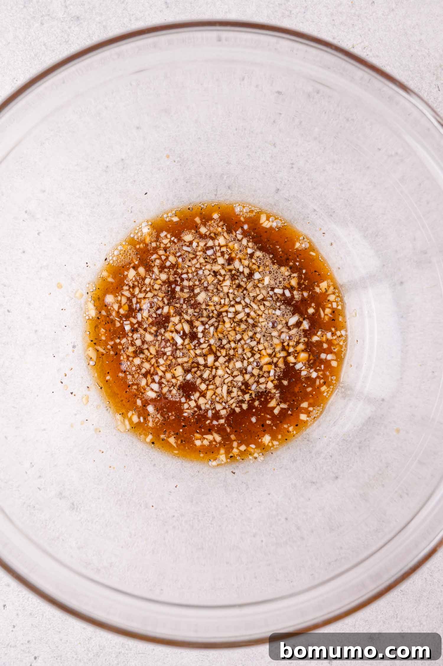 Crispy Honey Garlic Salmon Bites in the Air Fryer 7 A clear glass bowl containing a mixture of brown liquid with visible minced garlic and spices on a light countertop.