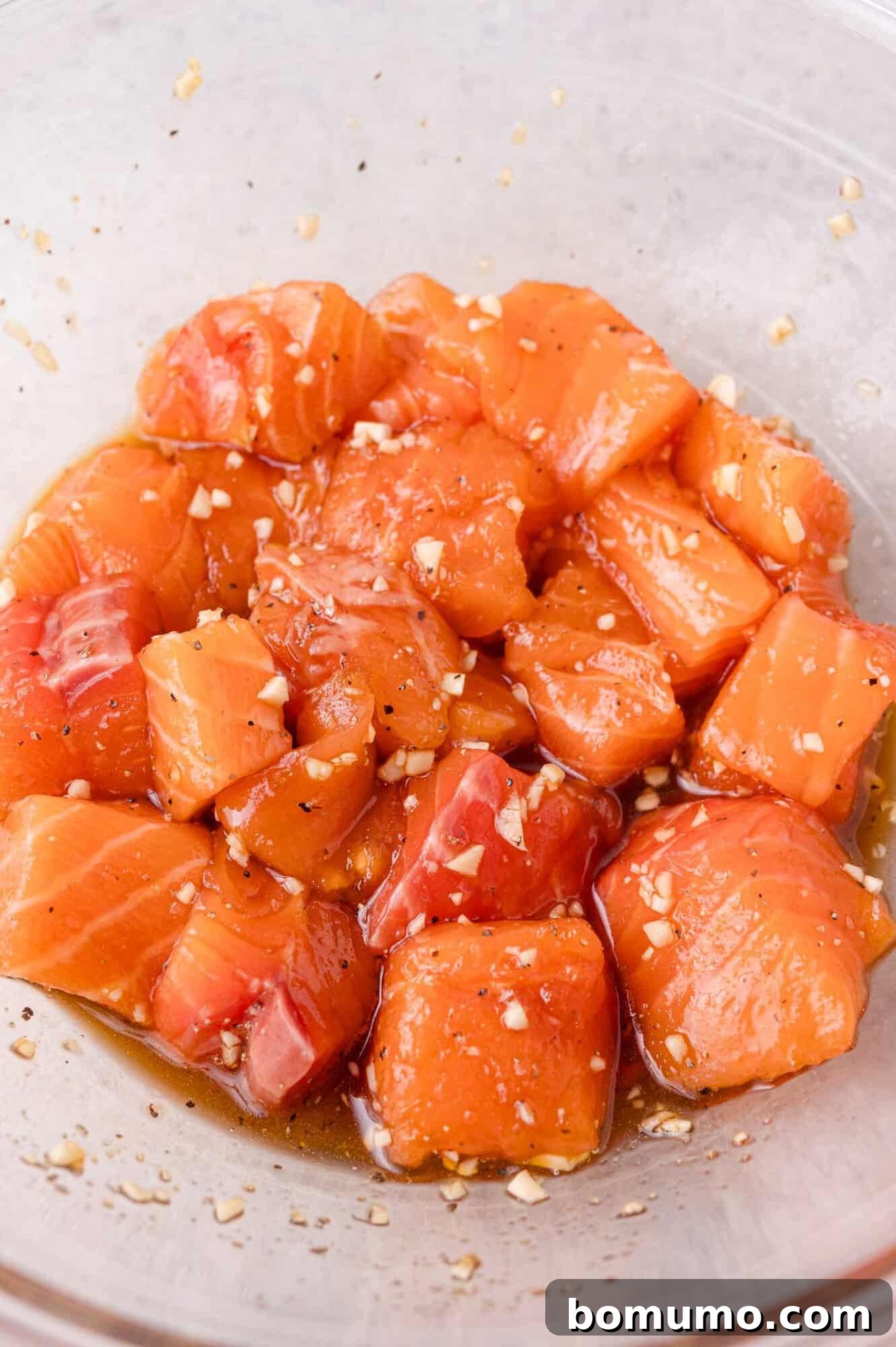 Crispy Honey Garlic Salmon Bites in the Air Fryer 8 Cubes of raw salmon marinated with sauce and minced garlic in a clear glass bowl.