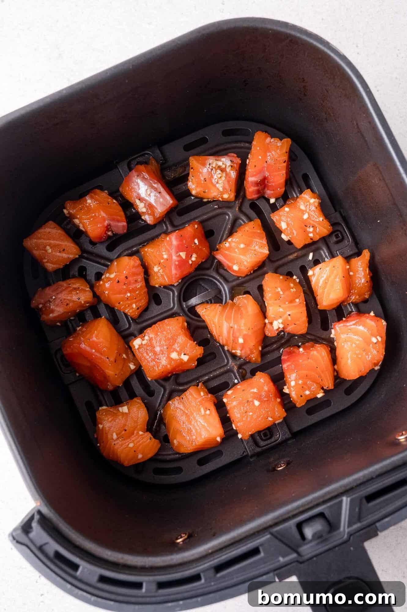 Crispy Honey Garlic Salmon Bites in the Air Fryer 9 Cubed raw salmon pieces, seasoned with spices, are arranged in a single layer inside an air fryer basket on a light-colored countertop.