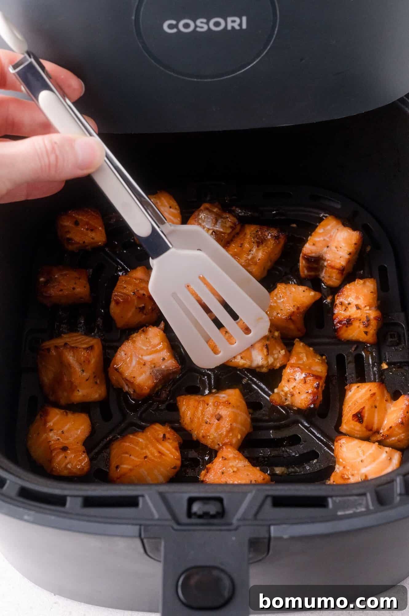 Crispy Honey Garlic Salmon Bites in the Air Fryer 10 Hand holding a spatula above cooked salmon bites inside an open Cosori air fryer basket.