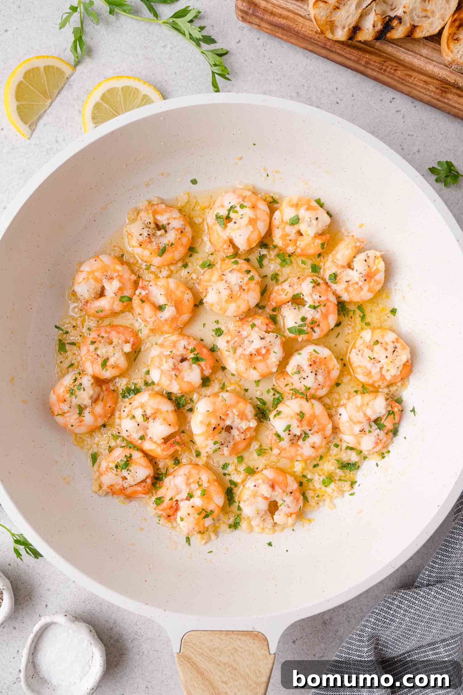 Garlic Butter Shrimp Scampi with Lemon and Parsley 2 Shrimp scampi cooked in a white skillet with garlic, herbs, and lemon butter sauce, garnished with parsley. Lemon slices and grilled bread are visible in the background.