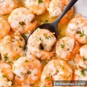Garlic Butter Shrimp Scampi with Lemon and Parsley 16 Cooked shrimp topped with minced garlic and chopped herbs, served in a garlic butter sauce with a black spoon scooping a piece.