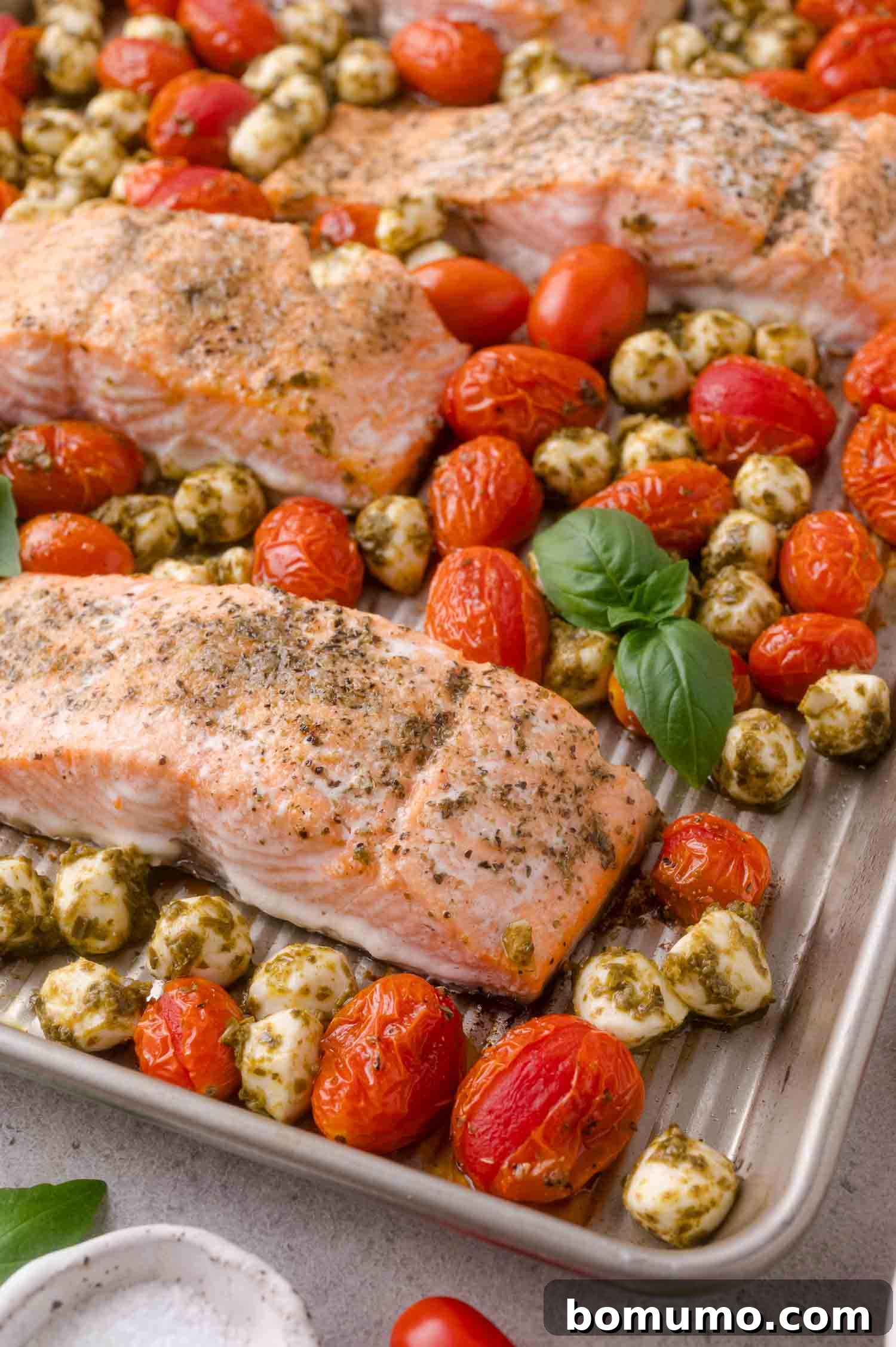Sheet Pan Caprese Salmon with Tomatoes and Basil 2 Baked salmon fillets on a sheet pan with roasted grape tomatoes, pesto-coated mozzarella balls, and fresh basil leaves.