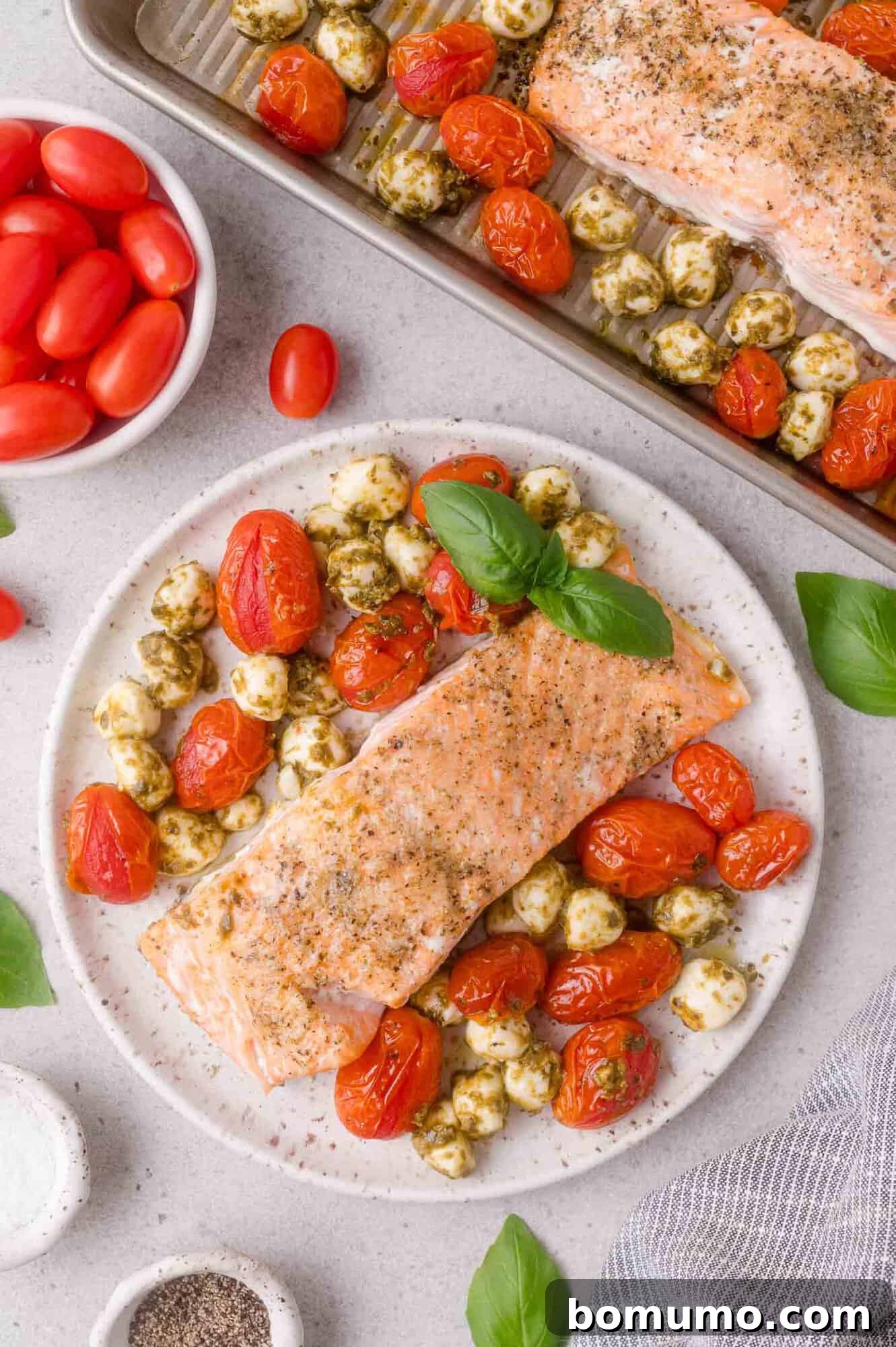 Sheet Pan Caprese Salmon with Tomatoes and Basil 12 Baked salmon fillet with roasted cherry tomatoes and mozzarella balls garnished with basil, served on a white plate next to a tray with more salmon and tomatoes.