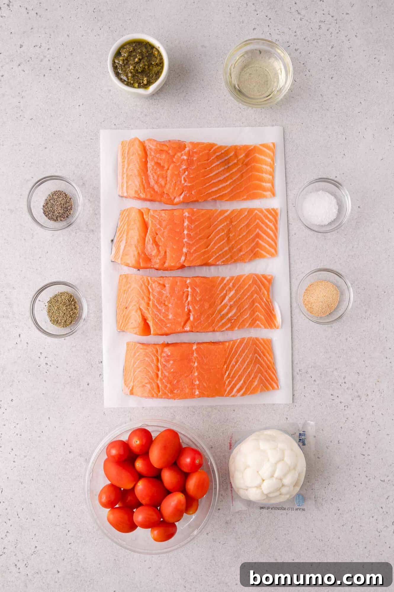 Sheet Pan Caprese Salmon with Tomatoes and Basil 3 Four raw salmon fillets on parchment paper, surrounded by bowls of grape tomatoes, mozzarella, seasonings, oil, and pesto on a gray countertop.
