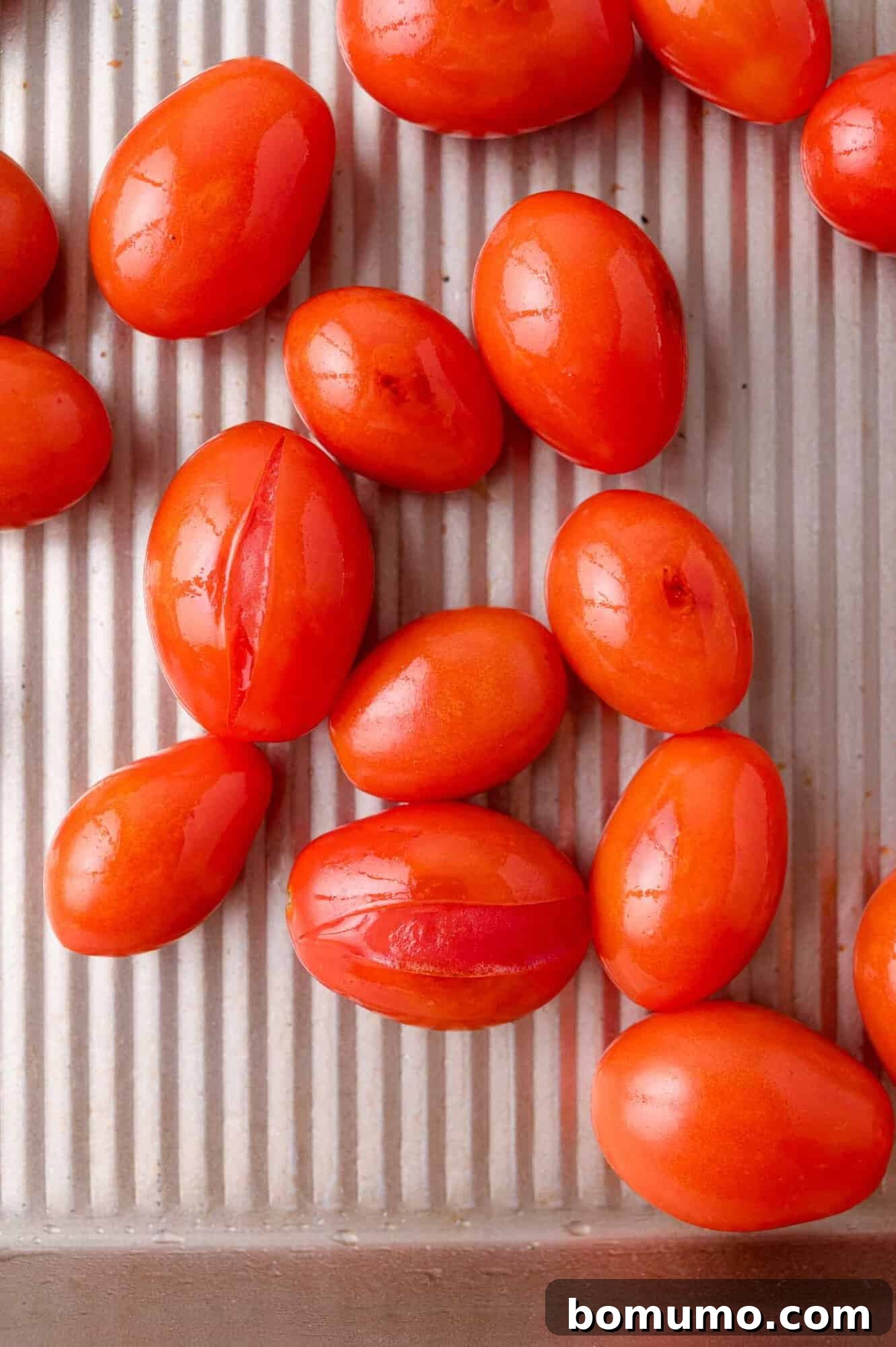 Sheet Pan Caprese Salmon with Tomatoes and Basil 6 Several oval-shaped, bright red tomatoes are arranged on a ridged metal baking tray, some with splits in their skins.