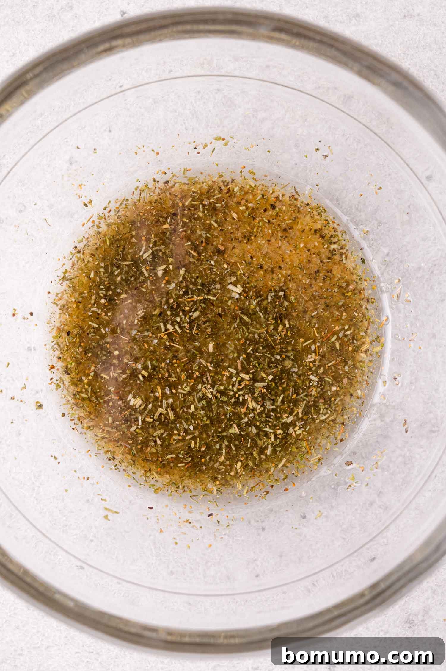 Sheet Pan Caprese Salmon with Tomatoes and Basil 7 Clear glass bowl containing a mixture of oil and dried herbs on a light surface.