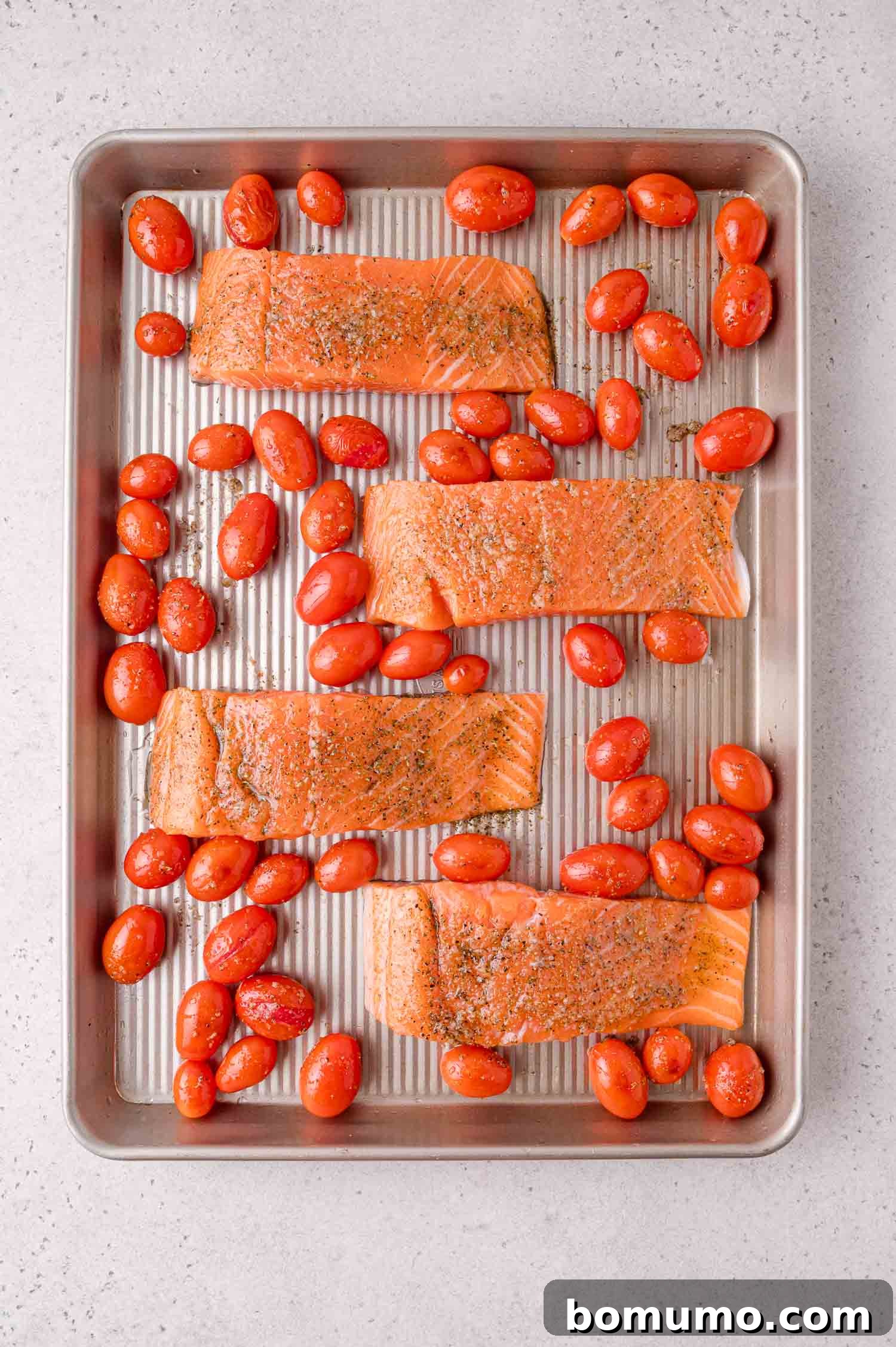 Sheet Pan Caprese Salmon with Tomatoes and Basil 8 Four raw salmon fillets and grape tomatoes are arranged on a metal baking sheet, seasoned with herbs and pepper, ready for roasting.