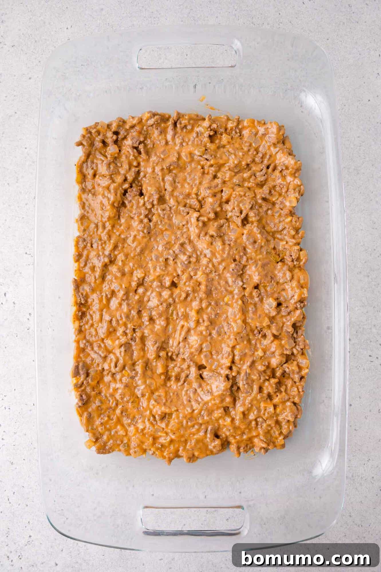 Cheeseburger Tater Tot Casserole Made Without Canned Soup 7 A glass baking dish filled with an even layer of ground beef mixed with a creamy sauce, placed on a light gray countertop.