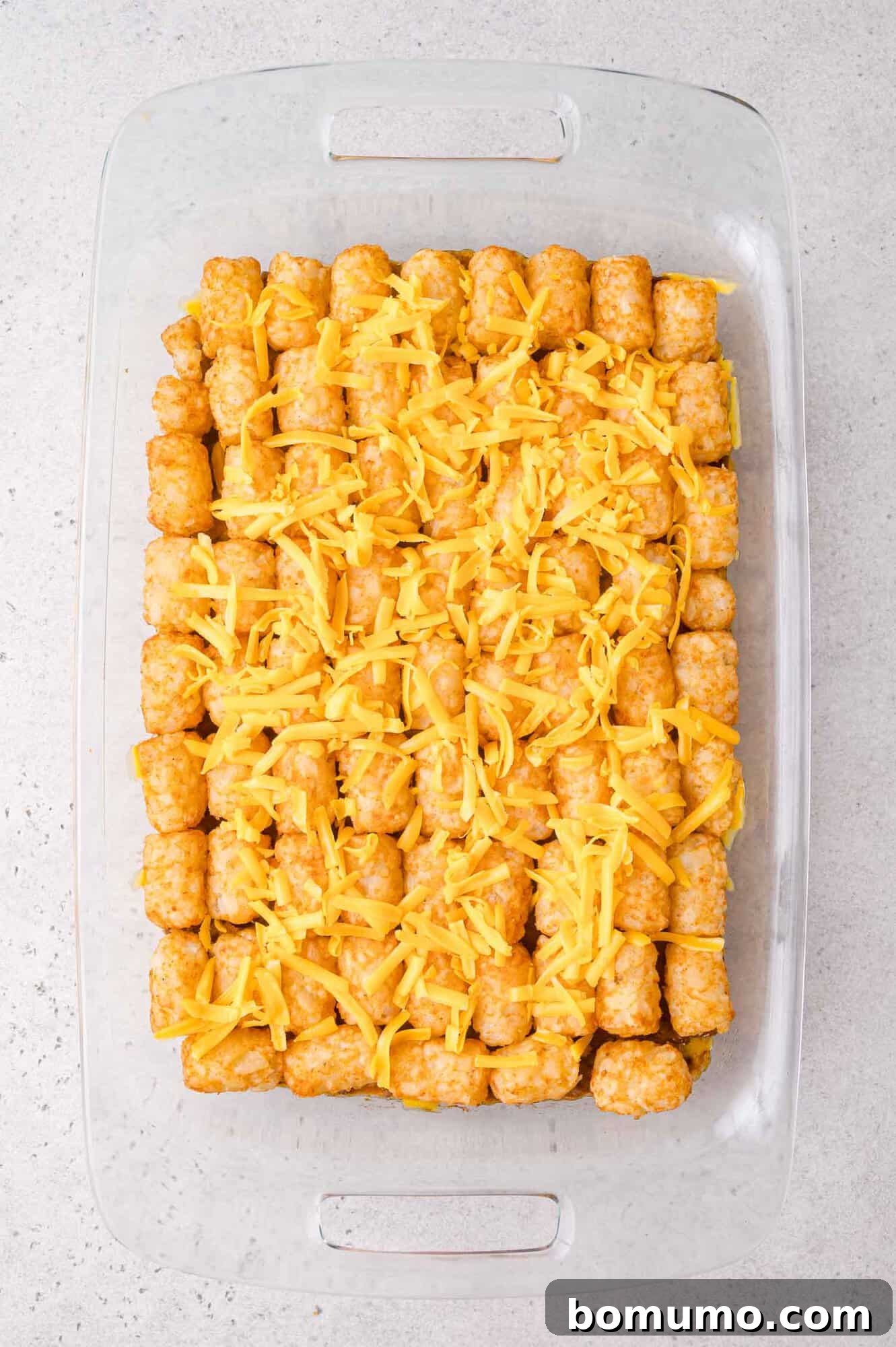 Cheeseburger Tater Tot Casserole Made Without Canned Soup 9 A glass baking dish filled with neatly arranged tater tots topped with shredded cheddar cheese on a light countertop.