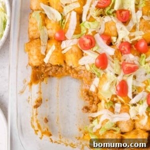 Cheeseburger Tater Tot Casserole Made Without Canned Soup 11 Glass baking dish with a portion of cheeseburger tater tot casserole topped with shredded lettuce, diced tomatoes, and melted cheese; one section is scooped out.