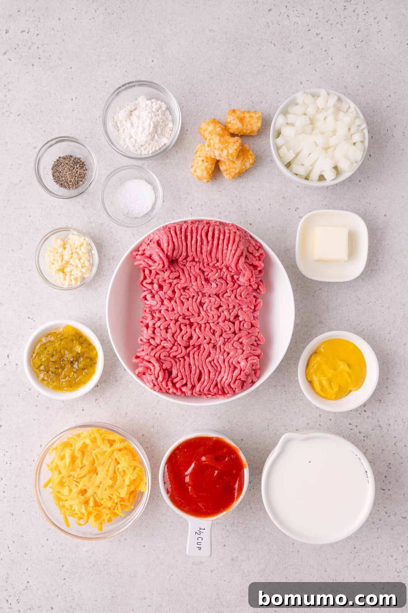 Cheeseburger Tater Tot Casserole Made Without Canned Soup 3 Ground beef, shredded cheese, diced onions, tater tots, milk, ketchup, mustard, relish, butter, flour, salt, pepper, and garlic arranged in bowls on a gray countertop.