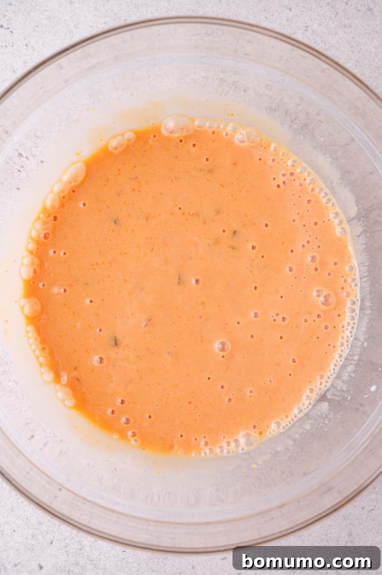 Cheeseburger Tater Tot Casserole Made Without Canned Soup 5 A clear glass bowl containing a frothy, orange mixture of the sauce ingredients with small bubbles on the surface, set on a light-colored countertop.