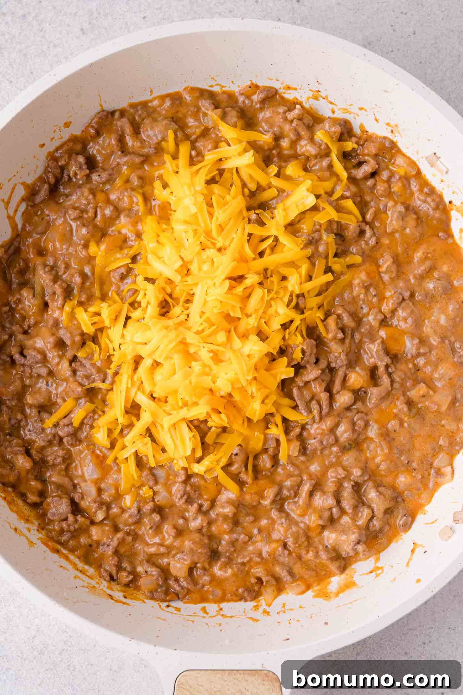 Cheeseburger Tater Tot Casserole Made Without Canned Soup 6 Ground beef in a creamy sauce is cooking in a white skillet, topped with a pile of shredded cheddar cheese.